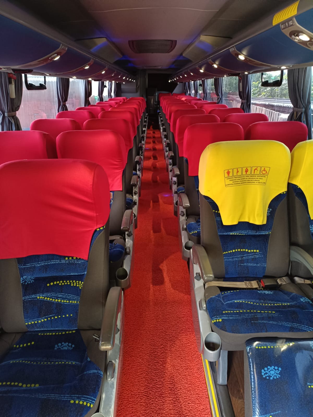 Interior do ônibus com corredor vermelho, bancos azuis e capas vermelhas e amarelas