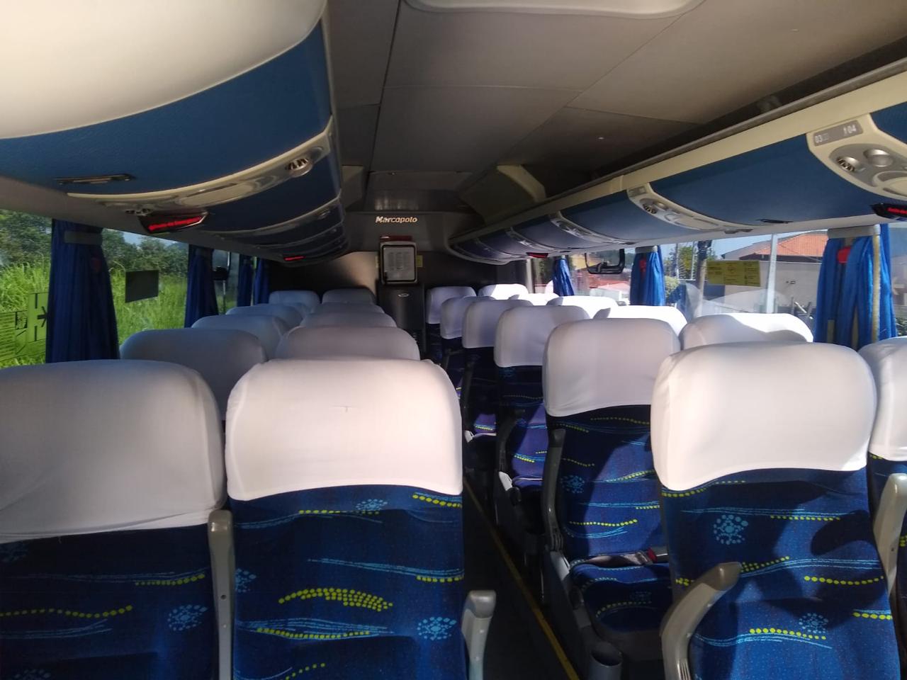 Interior do micro-ônibus com bancos azuis, capas brancas, corredor central e identificação Marcopolo